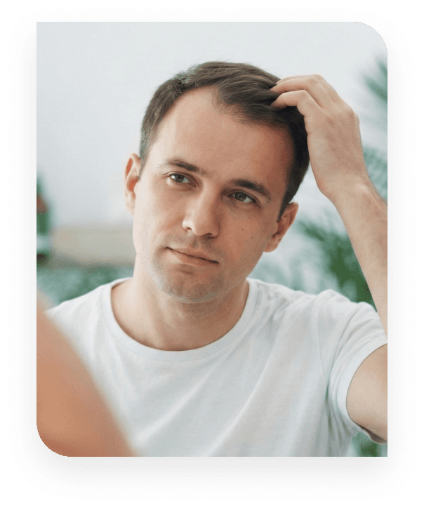 Man examining his hair and hairline in a mirror. A front view of a man in a white t-shirt looking at his reflection in a mirror while running his fingers through his hair, inspecting his hairline or scalp concern.